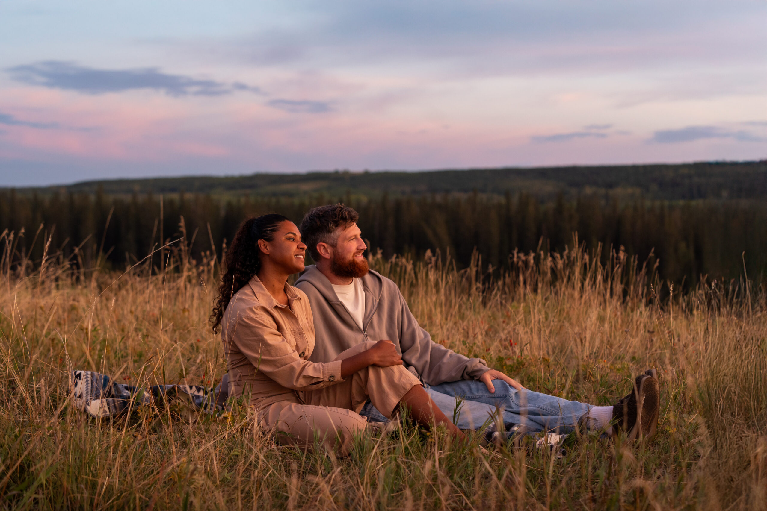 Woman and man sitting in a field of grass looking off at the horizon with the sun setting in the background