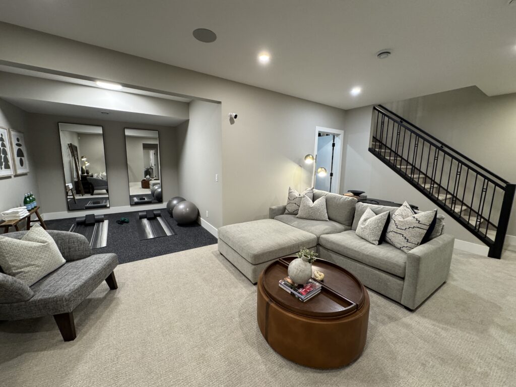Open basement with cream carpet, grey sectional, and alcove with two tall mirrors, yoga mats and free weights.