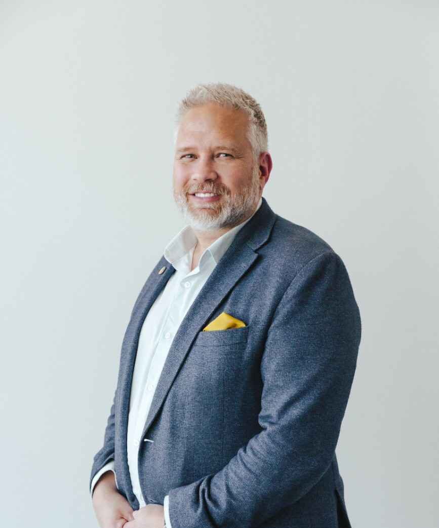 Man with light facial hair wearing white dress shirt and dark grey blazer with yellow pocket square smiling in front of white wall