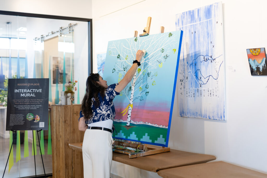 Woman stamping a leaf onto a colourful collaborative painting featuring a white birch tree.