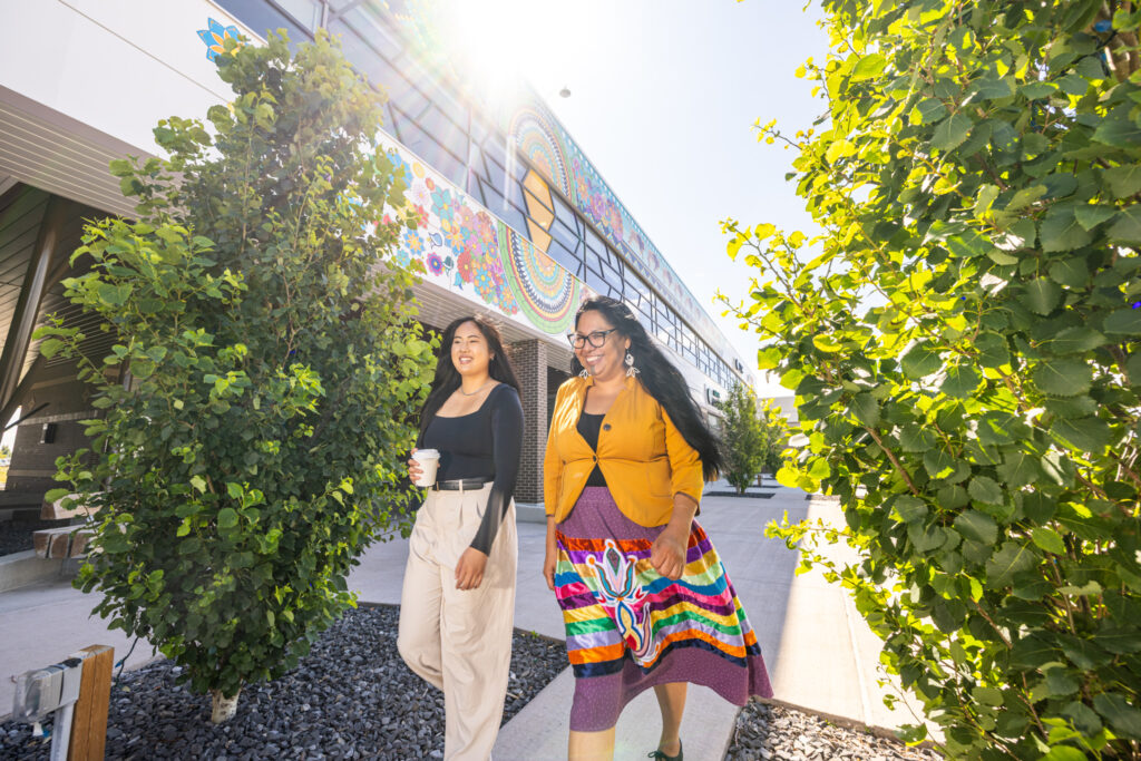 Two people walking through tree-lined sidewalks in The Shops at Buffalo Run with a colourful mural behind them.