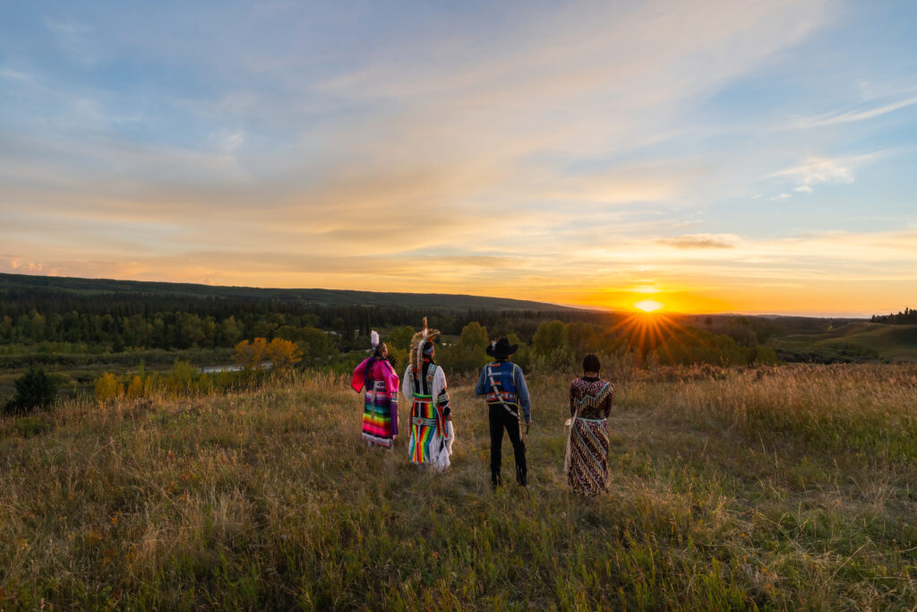 Group of Tsuut'ina Nation people in traditional regalia standing in a field watching the sun set.