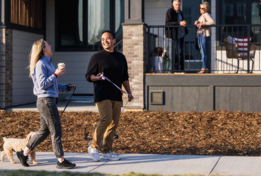 Man and woman each walking a small dog while chatting and laughing as they walk down the sidewalk. A couple stands on the sunny townhome porch in the background.