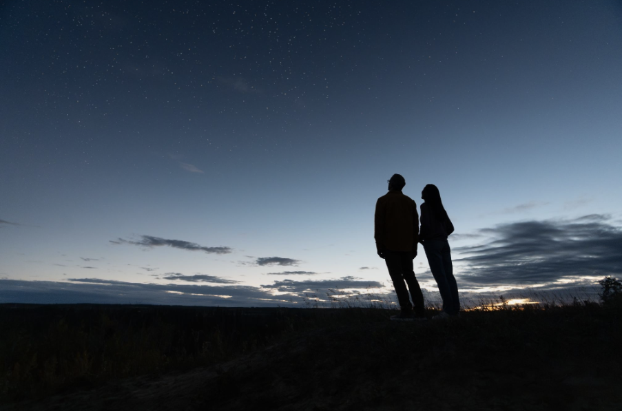 Dark silhouette of a couple stargazing as the sun sets below the prairie grass.