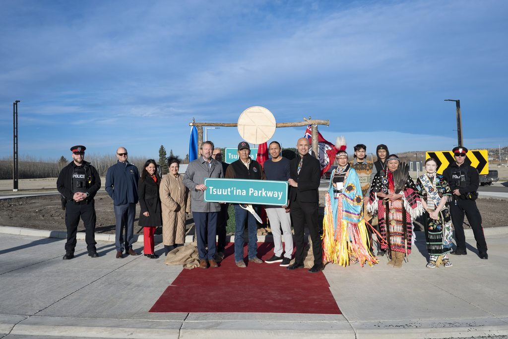 Tsuut’ina Nation leaders, Taza Development Corp. representatives, dancers in regalia and police officers stand on a red carpet at the Tsuut’ina Parkway roundabout, holding the road sign and ceremonial scissors.